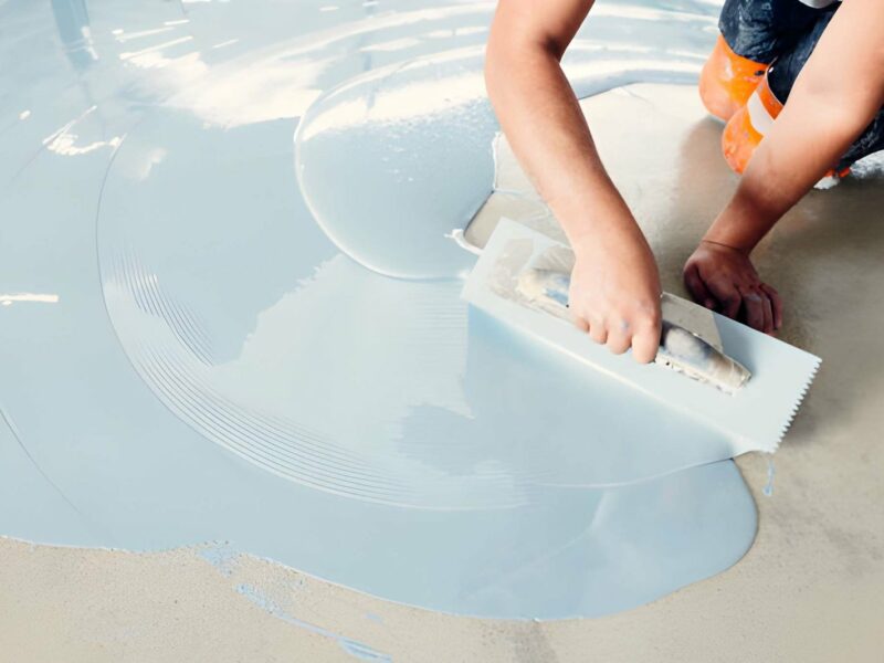 Flooring contractor using a notched trowel to spread light blue epoxy resin across a prepared concrete floor